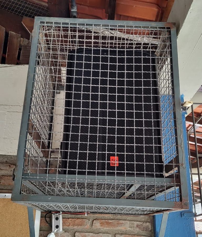 Caixa de som instalada com gaiola de proteção no pátio coberto de uma escola
